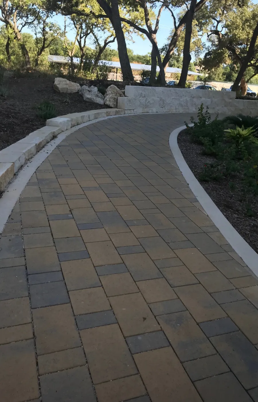 A curved paver walkway edged in limestone blocks, running between planted beds under live oaks